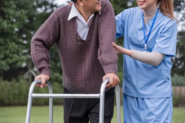 Asian female caregiver helps senior man walk A senior using a walker is assisted by a nurse at home. A nurse helps an elderly patient at a nursing home. Elderly care and healthy lifestyle.