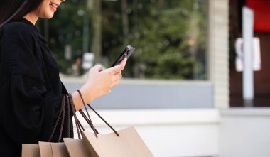 Akıllı telefon kullanan Asyalı kadın alışverişin tadını çıkarırken gözlerini kaçırıyor. Kara Cuma indirimi. Tatil için kıyafet almak..