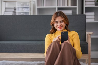 Mutlu Asyalı kadın evde koltukta otururken cep telefonuyla oynuyor. Genç kadın, sabah rahat bir şekilde kulaklıkla gülümsüyor. Dinlenme zamanı...