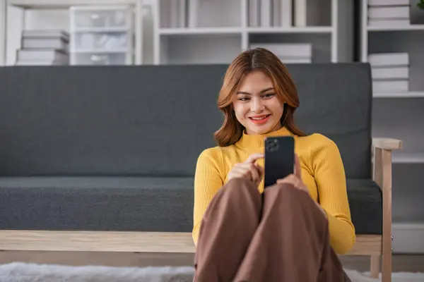 Happy Asian woman playing with mobile phone while sitting on sofa at home Young woman smiling relaxedly with headphones in the morning Its time to rest..