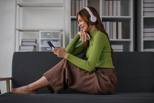 Happy Asian woman listening to music on mobile phone while sitting on sofa at home Relaxed smiling girl with headphones in the morning Its time to rest..