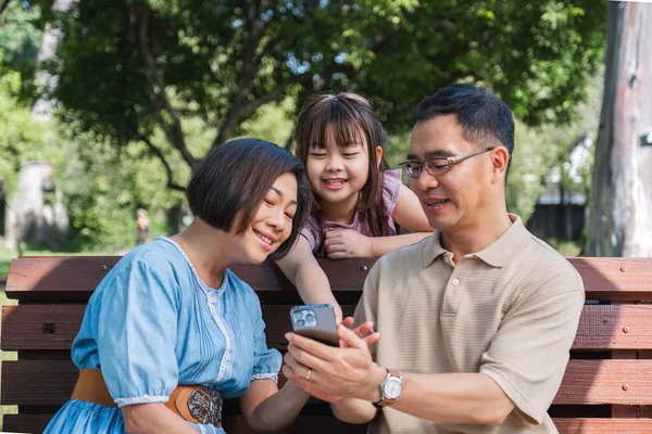 Kind and happy Asian senior grandparents enjoying looking at photos on their mobile phone in the park with their cute little granddaughter. On a clear day together lovely family.