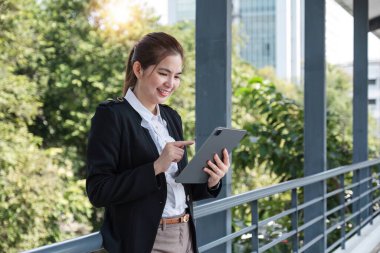 Asyalı iş kadını elinde tablet kontrol şirketinde çalışıyor. Tabletli kendine güvenen kadın çalışanı şehrin arka planında kameranın önünde duruyor..