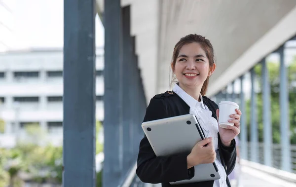 Asian businesswoman holding a laptop and a coffee mug. Smiling at the camera. Confident female employee holding a laptop standing in front of the camera on the background of a city skywalk..