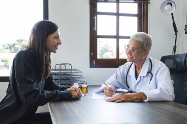 Beyaz önlüklü yaşlı kadın doktor, klinikte genç ve güzel bir hastayla konuşuyor, kalp hastalığı tedavisi ve sağlık ve tıp konsepti hakkında tavsiyelerde bulunuyor..