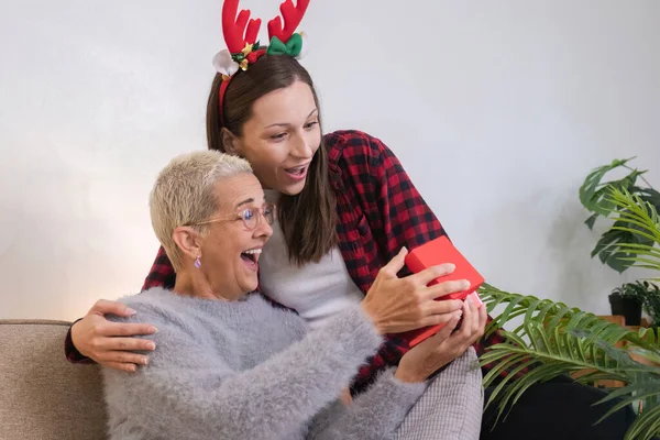 Sevimli genç bir kadın Noel günü annesine sürpriz yapmak için bir hediye kutusu tutuyor. 60 yaşında emekli bir kadın mutlu bir kız çocuğu Noel günü oturma odasında elinde kırmızı bir hediye kutusu tutuyor..