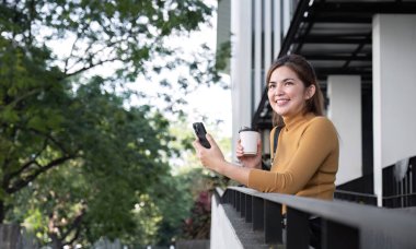 Başarılı Asyalı iş kadını ayakta duruyor ve gülümsüyor. Cep telefonunuzdaki uygulamayı kullanarak şehirde kalın. Akıllı telefonunuzdaki hızlı bağlantıdan haberleri okuyun Açık hava mobil uygulamasına bakın.