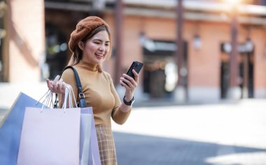 Güzel Asyalı kadın alışveriş torbaları tutuyor, akıllı telefon kullanıyor ve Kara Cuma günü alışveriş merkezinin dışında alışveriş yaparken gülümsüyor...