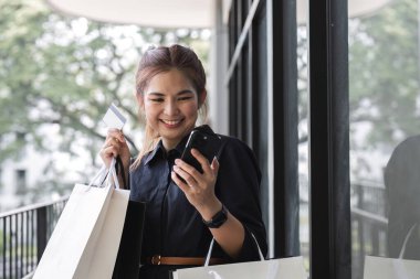 Akıllı telefon kullanan Asyalı kadın alışverişin tadını çıkarırken gözlerini kaçırıyor. Kara Cuma indirimi. Tatil için kıyafet almak..