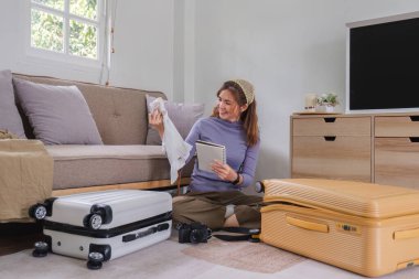 Güzel genç Asyalı kadın yurt dışına çıkmadan önce kıyafet ve ekipman hazırlamak için listeyi kullanıyor...