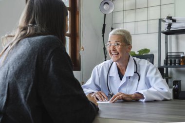 Beyaz önlüklü yaşlı kadın doktor, klinikte genç ve güzel bir hastayla konuşuyor, kalp hastalığı tedavisi ve sağlık ve tıp konsepti hakkında tavsiyelerde bulunuyor..