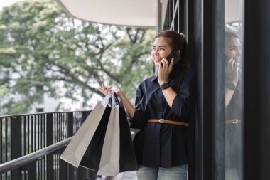 Akıllı telefon kullanan Asyalı kadın alışverişin tadını çıkarırken gözlerini kaçırıyor. Kara Cuma indirimi. Tatil için kıyafet almak..