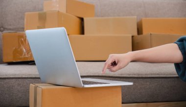 Çevrimiçi satışlarla ilgilenen genç bir kadın, küçük işletme girişimcisi, ürün siparişlerini kontrol etmek ve teslimata hazırlanmak için dizüstü bilgisayar kullanıyor...