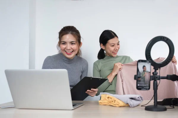 Young Asian female friends talk to a live camera on the phone to sell clothes together at home. and display clothing to customers in online sales.