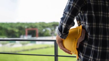 Male engineer standing holding safety helmet looking out at construction site.. clipart