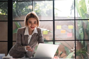 Gülümseyen Asyalı iş kadını finansal işlemler, vergi hesapları, cam ofisindeki çalışma masasında planlama yapmak için dizüstü bilgisayar kullanıyor...