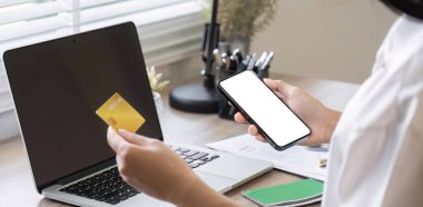 Elinde kredi kartı olan genç bir kadın online alışveriş için dizüstü bilgisayar kullanıyor ve akıllı telefondan internet üzerinden ödeme yapıyor..