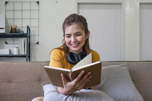 Young Asian woman sitting on modern sofa in front of window, relaxing in living room, reading a book at home..
