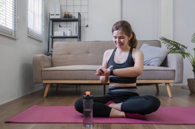 Genç, formda bir Asyalı kadın oturma odasında yerde oturuyor ve akıllı saatine bakıyor. Sporcunun biri spor yaptıktan sonra spor programında performansını izliyor...