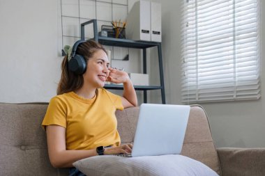 Asyalı bir kadın oturma odasında dizüstü bilgisayar kullanarak ve kulaklıkla müzik dinleyerek oturuyor...