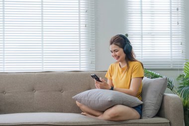 Mutlu Asyalı kadın evdeki koltukta oturmuş cep telefonundan müzik dinliyor, sabah kulaklıkla gülümseyen kız, dinlenme zamanı...