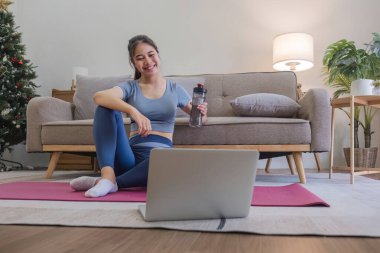 Genç Asyalı kadın evde yoga yaparken dizüstü bilgisayar kullanıyor ve su içiyor. Rahatlama zamanı.