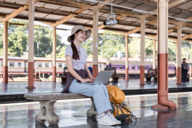 Modern tren istasyonunda genç bir Asyalı kadın. Sırt çantası yolcusu. Tren beklerken dizüstü bilgisayar kullanıyor.