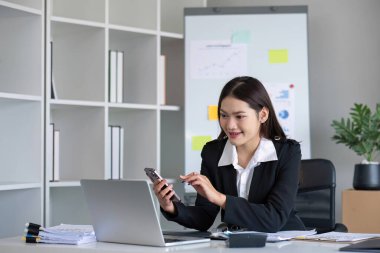 Mutlu Asyalı iş kadını modern ofiste akıllı telefon, tablet ve dizüstü bilgisayarla konuşma zevkine sahip..