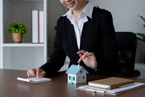 Female real estate agent is calculating budget before signing real estate project contract with house model at desk..