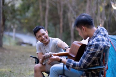 Asyalı LGBTQ çifti doğanın tadını çıkarıyor, nehir kenarındaki orman bölgesinde çadır kuruyorlar, gitar çalıyorlar...