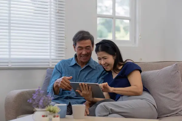 Cheerful Asian couple using network video call tablet sitting together on sofa at home Asian elderly husband sitting with elderly wife shopping online or video calling on tablet, relaxing..