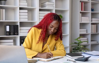 Genç Afrikalı Amerikalı iş kadını rakamları hesaplamak için bir hesap makinesi kullanıyor. Şirketin mali hesapları ofisteki masada..