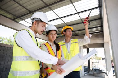 Profesyonel inşaat mühendisleri ve mimarlardan oluşan bir ekip proje planı kağıtlarını tutuyor, inşaat alanındaki inşaat projelerini denetliyor ve kontrol ediyor...