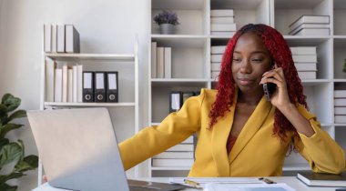 Genç Afrikalı Amerikalı iş kadını telefonda konuşuyor ve ofiste çalışırken notlar alıyor..