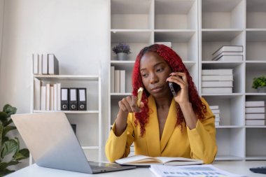 Genç Afrikalı Amerikalı iş kadını telefonda konuşuyor ve ofiste çalışırken notlar alıyor..