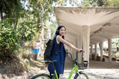 Genç Asyalı bir kadın işe giderken yeşil bir şehirde bisiklet sürüyor, sırt çantası taşıyor ve çevreye zarar vermemek için yeniden kullanılabilir bir bardak kullanıyor...