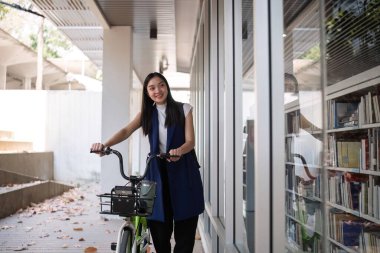 Genç Asyalı bir kadın işe giderken yeşil bir şehirde bisiklet sürüyor, sırt çantası taşıyor ve çevreye zarar vermemek için yeniden kullanılabilir bir bardak kullanıyor...