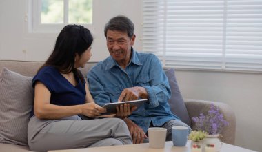 Neşeli Asyalı çift, evde, Asyalı yaşlı kocanın kanepede birlikte oturup, yaşlı eşiyle internetten alışveriş yapan veya tablet üzerinden video izleyen, rahatlatıcı bir video kullanıyor...