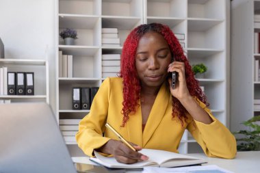 Genç Afrikalı Amerikalı iş kadını telefonda konuşuyor ve ofiste çalışırken notlar alıyor..