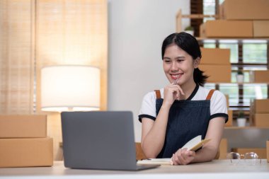 Genç bayan girişimci ürünleri çevrimiçi satmak için bir ürün kutusunun önüne yazıyor ve bunları müşterilere göndermeye hazırlanıyor. Evde satılıyor, evden çalışıyor..