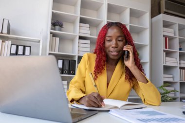 Genç Afrikalı Amerikalı iş kadını telefonda konuşuyor ve ofiste çalışırken notlar alıyor..