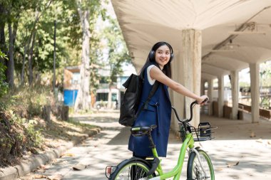 Genç Asyalı bir kadın işe giderken yeşil bir şehirde bisiklet sürüyor, sırt çantası taşıyor ve çevreye zarar vermemek için yeniden kullanılabilir bir bardak kullanıyor...