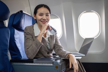 Kredi kartı alışverişini internet üzerinden yapan genç Asyalı kadın uçuş, seyahat ve iş konsepti sırasında uçağın birinci sınıf penceresinin yanında oturan dizüstü bilgisayarını kullanıyor...