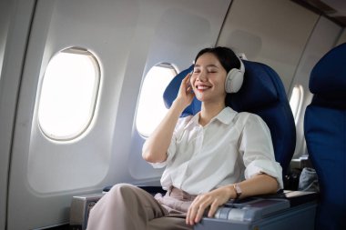 Seyahat sırasında kulaklık takan, müzik dinleyen, birinci sınıfta pencere kenarında oturan, uçuş, seyahat ve iş konsepti olan genç bir kadın..