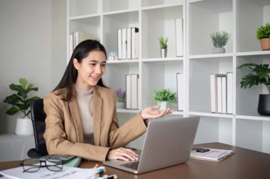 Genç Asyalı iş kadını karanlık yeşil bitkilerle süslenmiş modern bir ofiste dizüstü bilgisayar kullanarak oturur ve çalışır...
