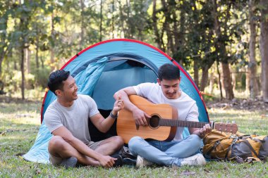 Asyalı LGBTQ çifti doğanın tadını çıkarıyor, nehir kenarındaki orman bölgesinde çadır kuruyorlar, gitar çalıyorlar...
