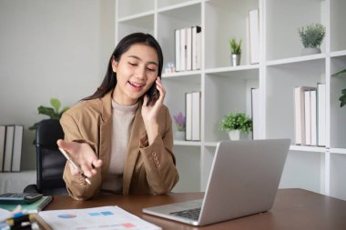 Genç Asyalı iş kadını, internet üzerinden yapılan bir iş toplantısında gölgeli yeşil bitkilerle süslenmiş modern bir ev ofisinde dizüstü bilgisayar kullanarak telefonda oturuyor...