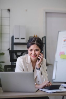 Genç Asyalı iş kadını, internet üzerinden yapılan bir iş toplantısında gölgeli yeşil bitkilerle süslenmiş modern bir ev ofisinde dizüstü bilgisayar kullanarak telefonda oturuyor...