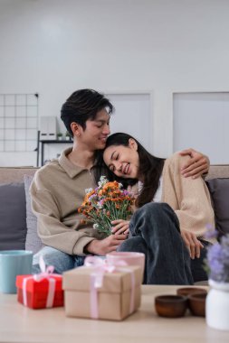 Genç çift sarılıyor ve birbirlerine sevgilerini gösteriyorlar. Özel günlerde ya da Sevgililer Günü 'nde birbirlerine çiçek buketleri verirlerdi. Oturma odasındaki kanepede..