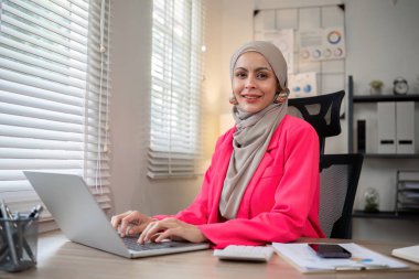Başörtüsü takan genç Müslüman girişimci özel ofiste dizüstü bilgisayarla çalışıyor...
